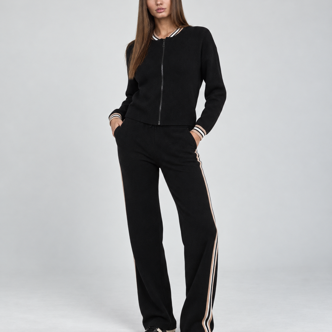 A woman modeling a black knit zip-up jacket and matching wide-leg pants with beige and white striped accents on the collar, cuffs, and sides, photographed against a light gray studio background.