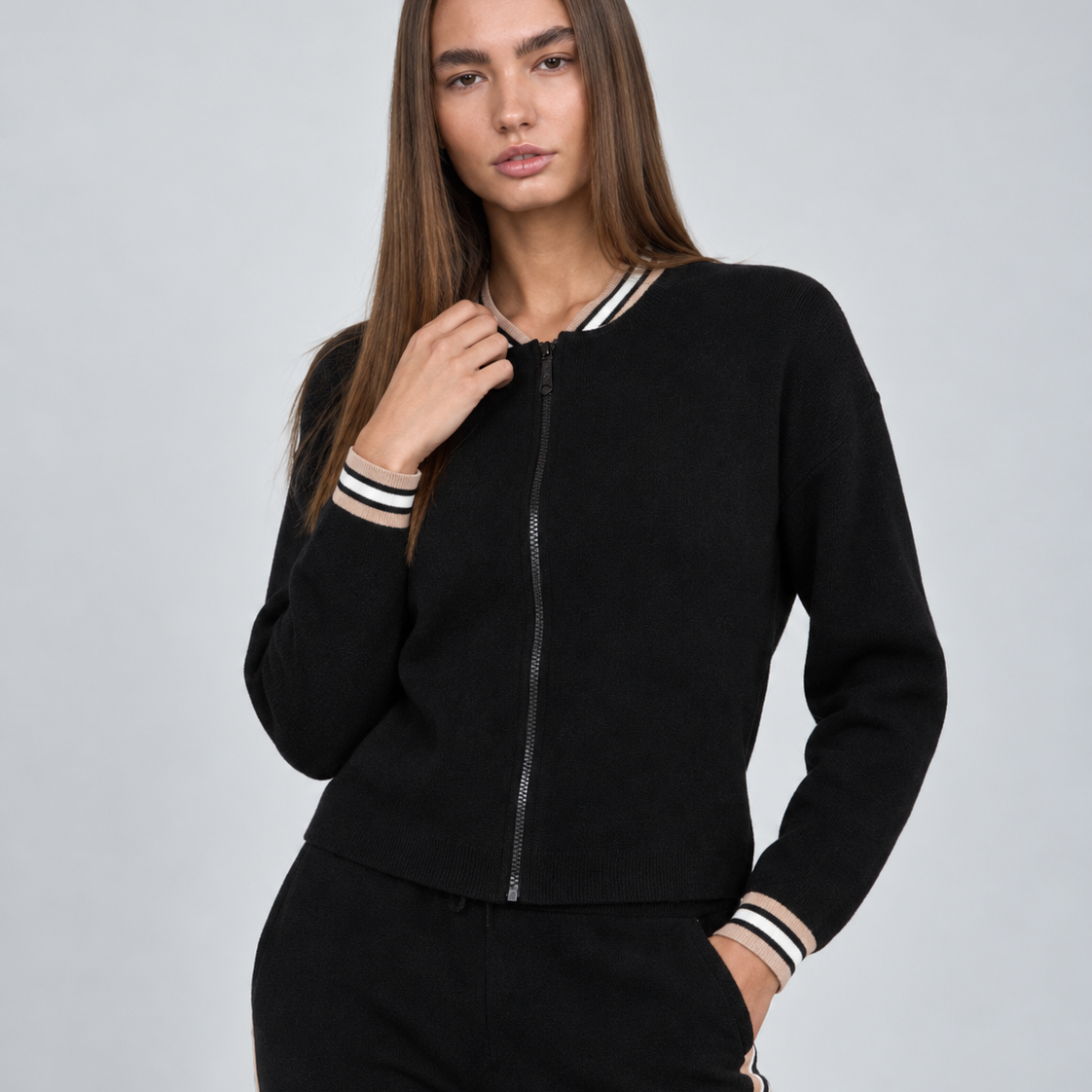 A woman modeling a black zip-up knit jacket with striped ribbed collar, cuffs, and hem, paired with matching black pants, against a light gray studio background.