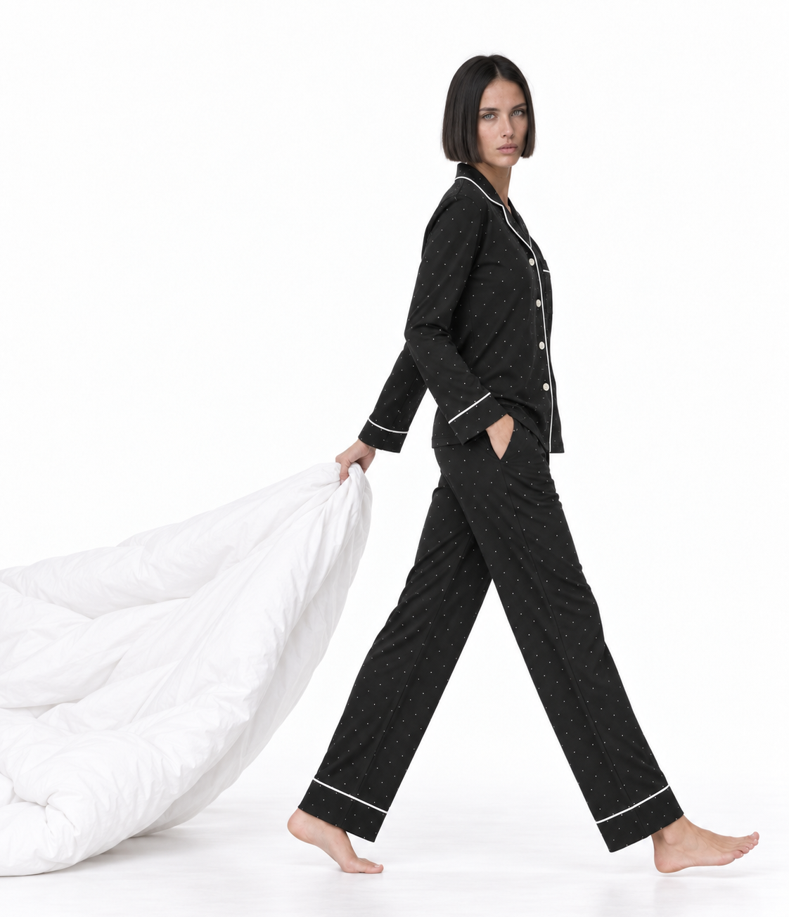A woman modeling a black two-piece pajama set with white piping, walking barefoot while holding a white duvet against a clean white studio background.