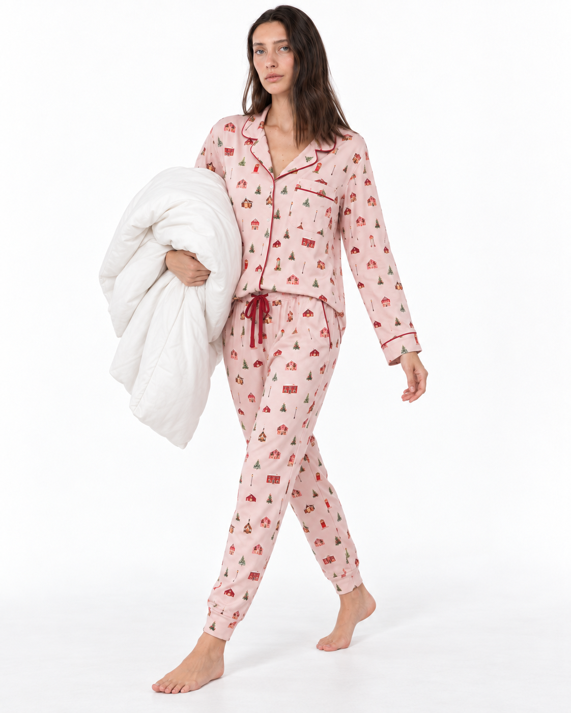 A woman modeling a light pink pajama set with a red Christmas tree and cabin pattern, holding a white quilted blanket against a plain white studio background.