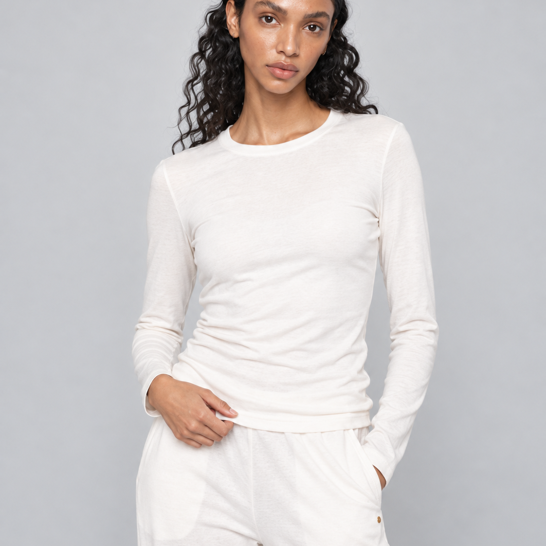 A woman modeling a matching off-white long-sleeve crewneck top and relaxed-fit pants against a solid gray studio background.