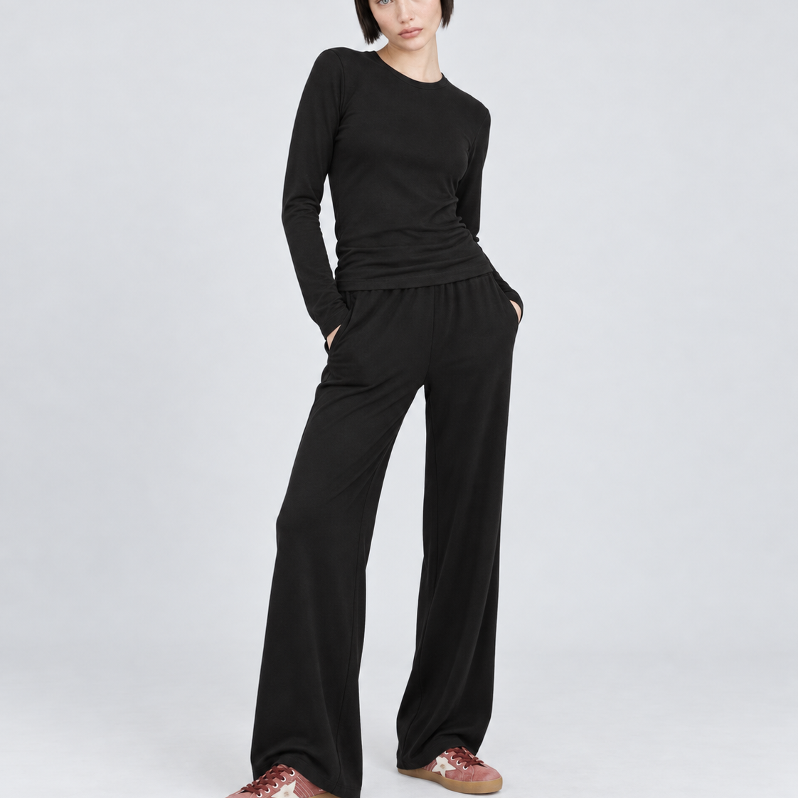 A model wearing a black long-sleeve top and matching black wide-leg pants, paired with brown slip-on shoes with white star details, standing against a light gray studio background.