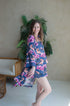 A woman standing in a room with a plant in the background, wearing a blue and multicolored floral lace-trimmed 3/4 sleeve robe.