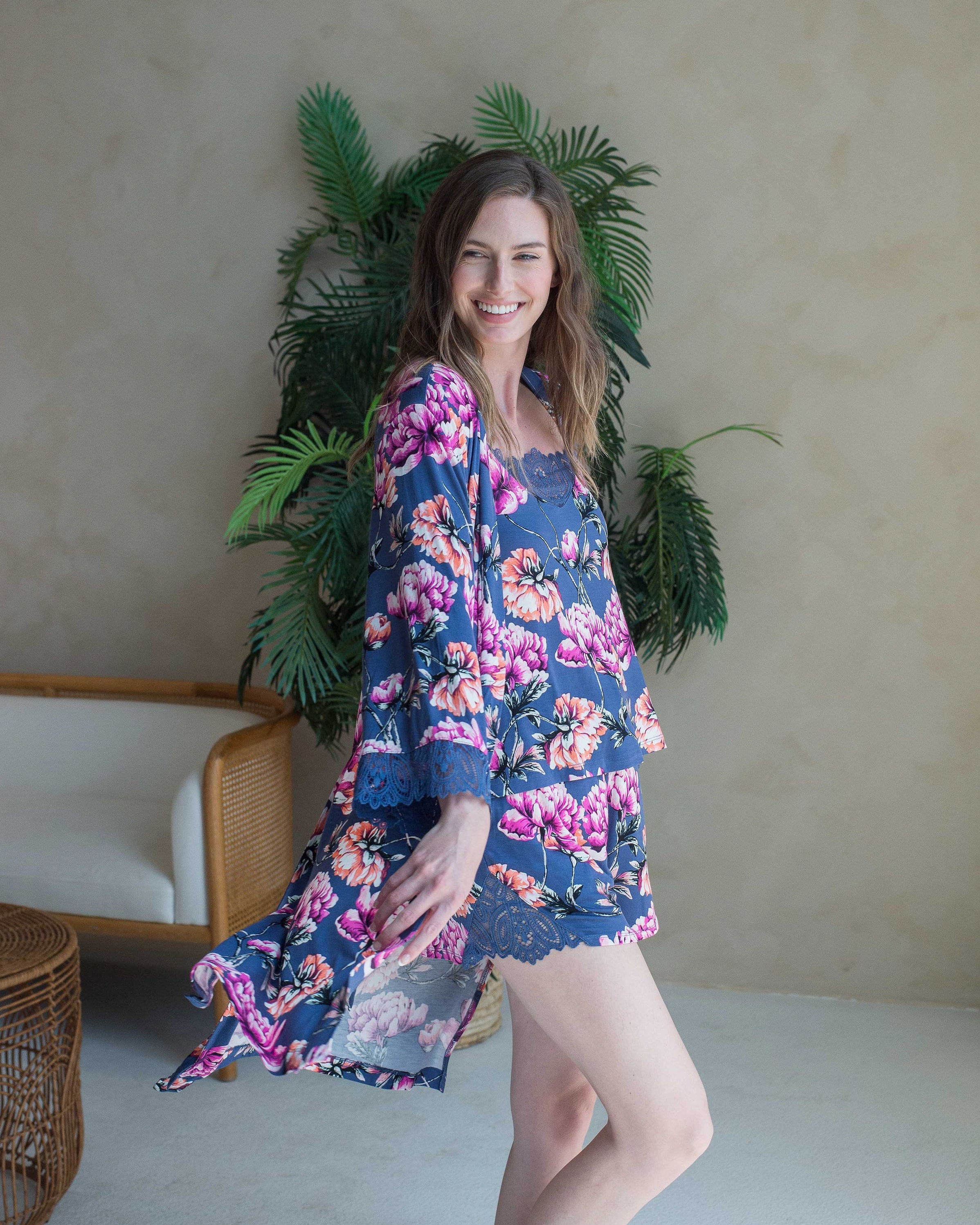 A woman standing in a room with a plant in the background, wearing a blue and multicolored floral lace-trimmed 3/4 sleeve robe.