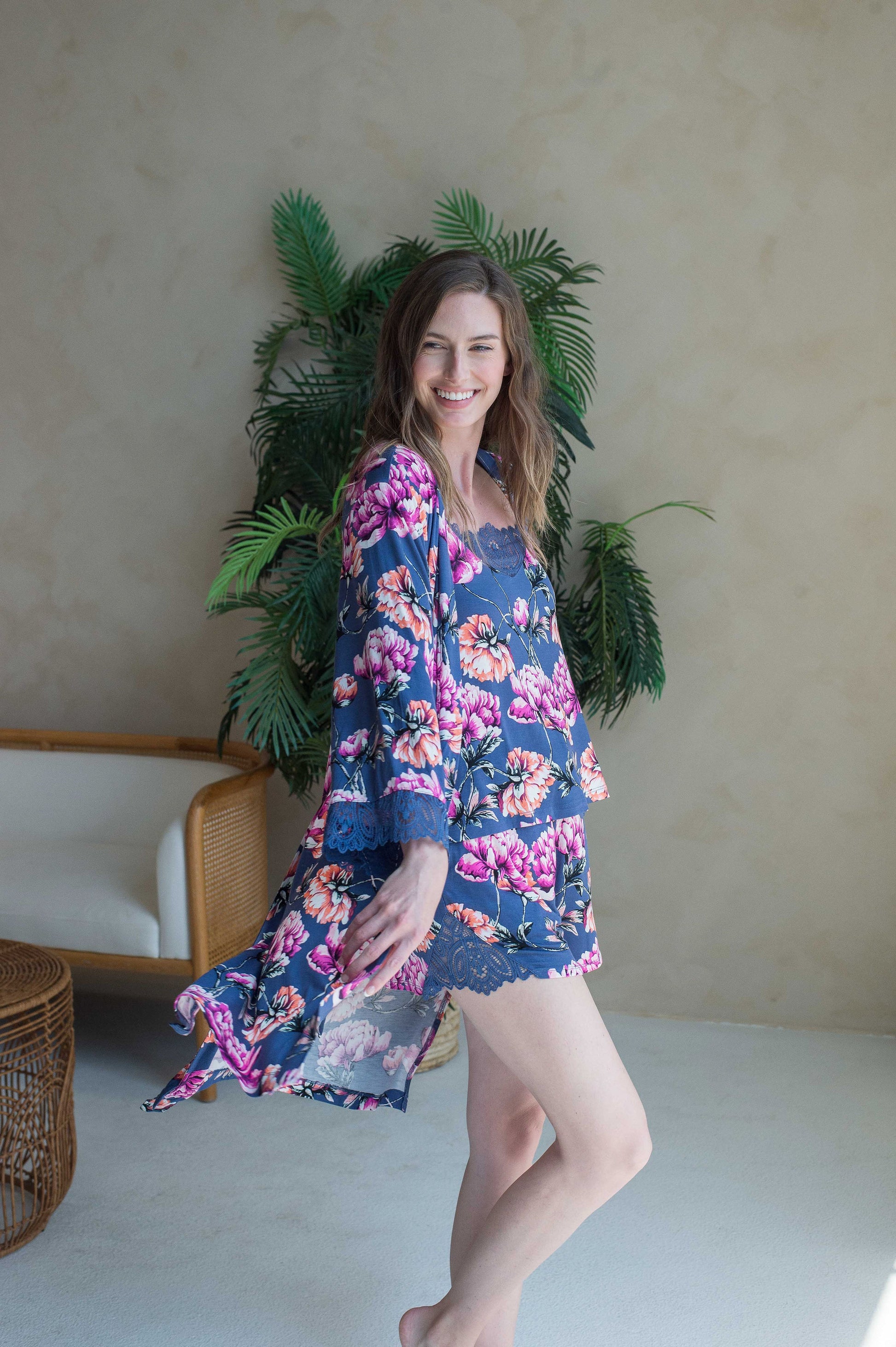 A woman standing in a room with a plant in the background, wearing a blue and multicolored floral lace-trimmed 3/4 sleeve robe.