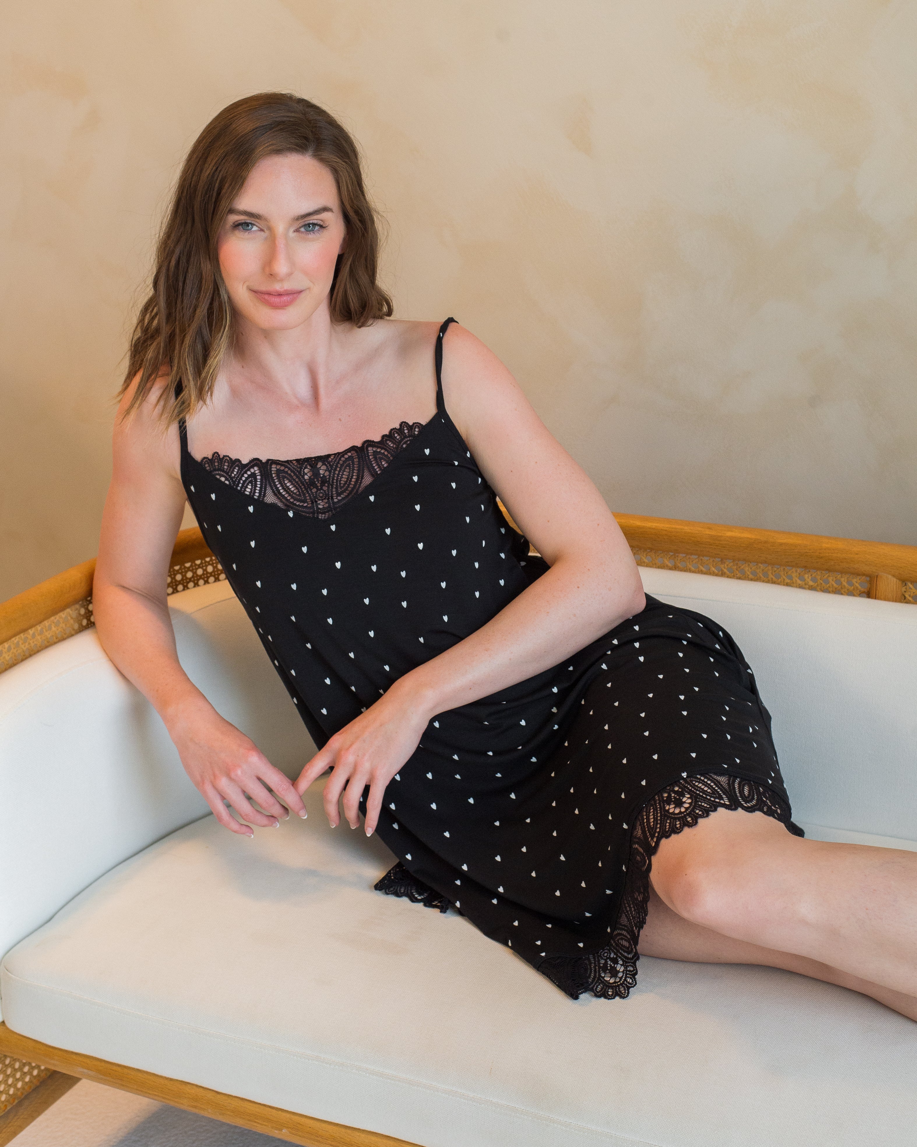 A woman sitting on a couch, wearing a black sleeveless lace chemise with lace trim on the neckline and hem, paired with a black strap crop top.