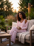 Woman in pink outfit sitting on a couch outdoors with plants and lights in the background